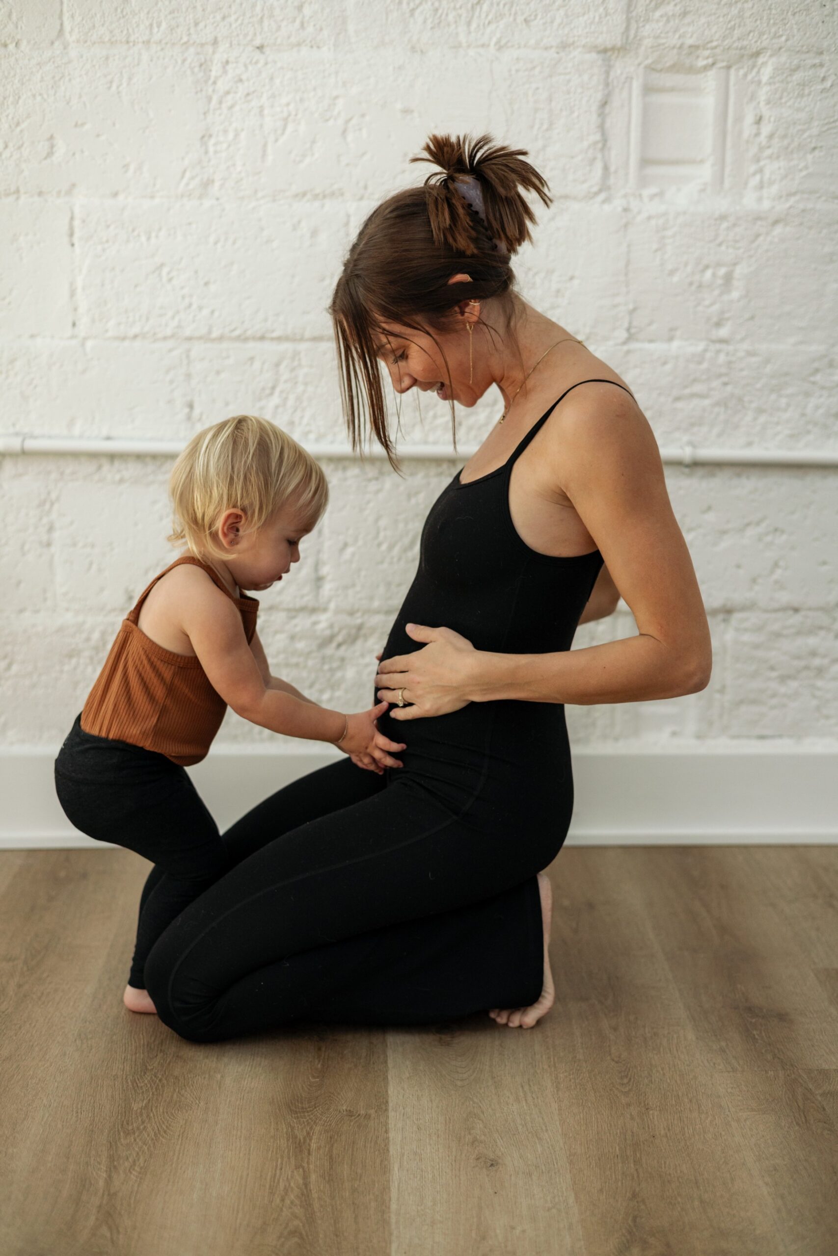 Pediatric aged baby on the left, in black leggings, and light brown tank top, looking at mom's belly with hands on her belly, and woman in black leggings and tank top with hands on belly smiling at pediatric girl.
