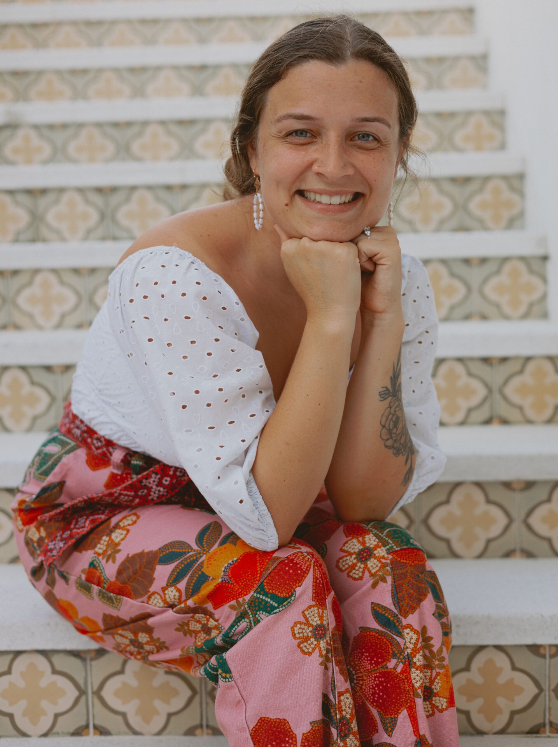 Woman smiling at camera, sitting on stairs, in a white shirt, and pink and red flower pants.