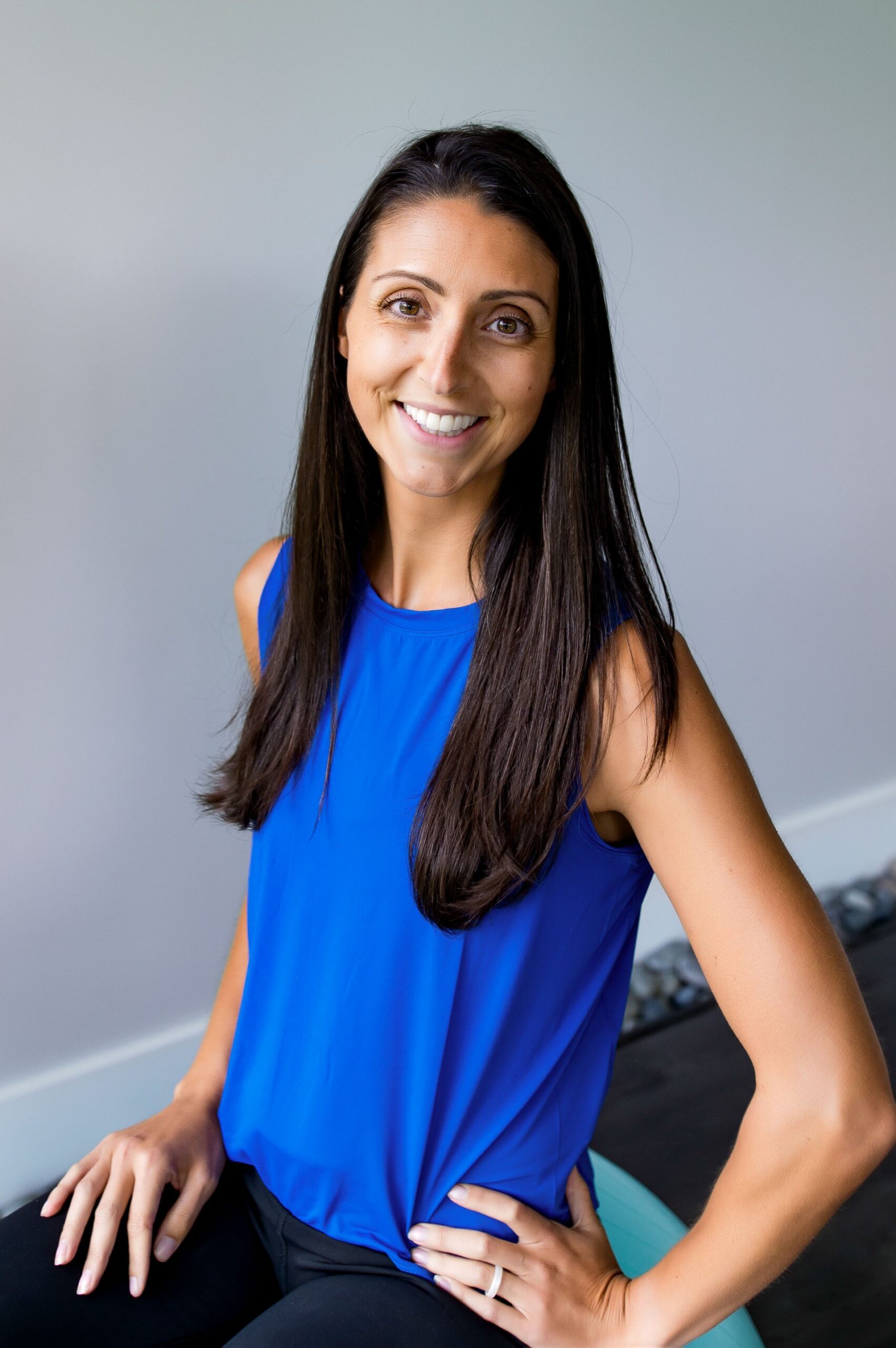 Woman smiling at camera with dark brown hair wearing a blue shirt and black pants, with left arm bent and hand on hip. and left hand resting on thigh