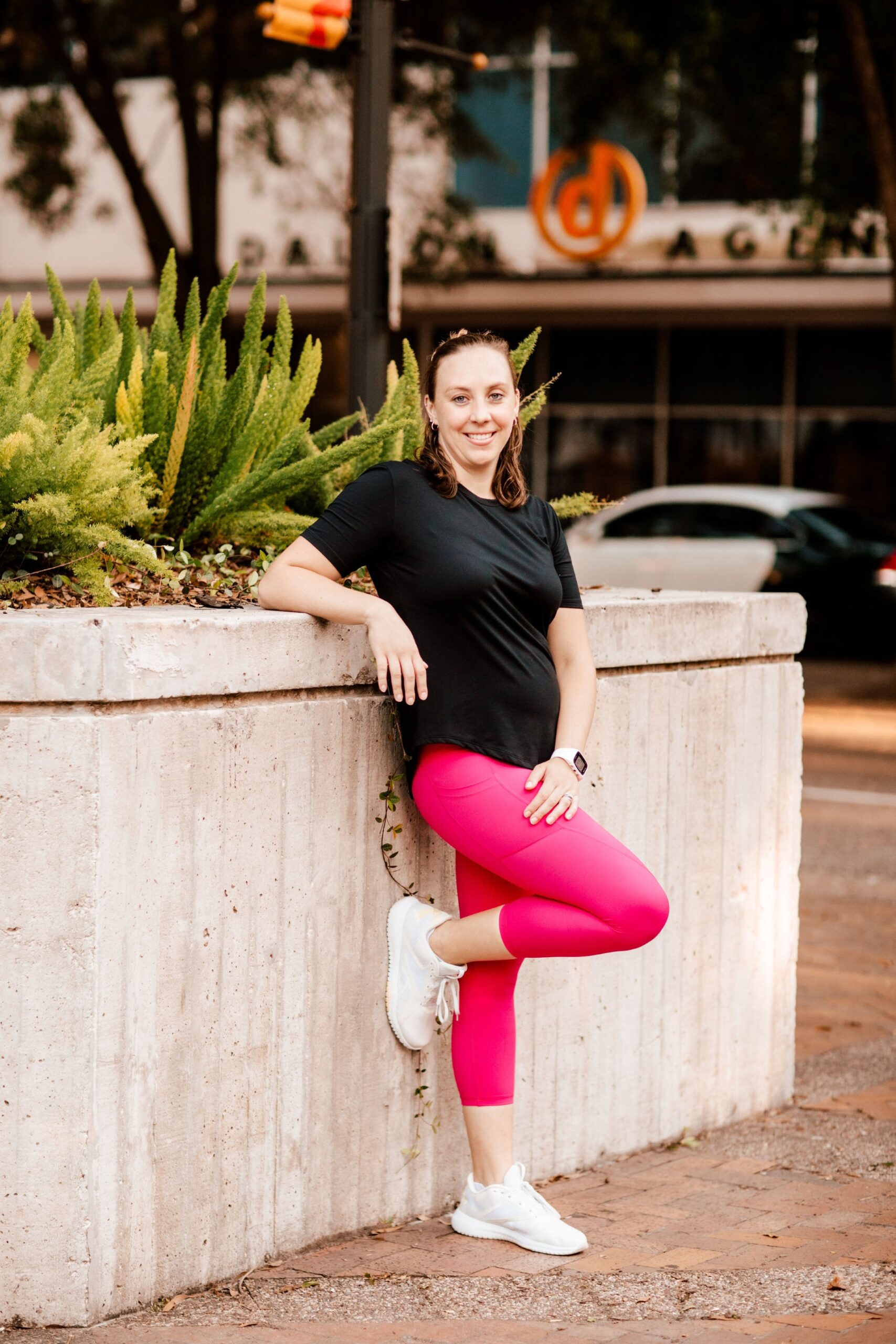 Light skinned woman in black shirt and pink pants, smiling at camera, leaning against cement piece with shrubs in it, in Jacksonville, FL