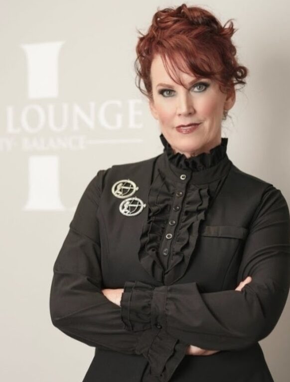 Light skinned woman standing with arms crossed, wearing a blakc shirt with two pins, red hair, and ZL, Zen lounge sign behind her.