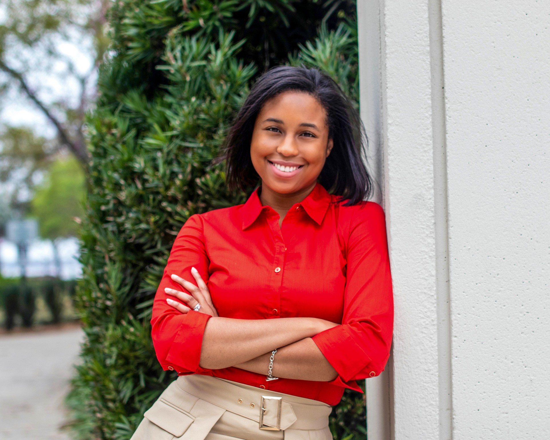 African American woman leaning against a building, smiling, wearing a red shirt and khaki pants, with her arms crossed, and green shrubs in the background, in Jacksonville, FL