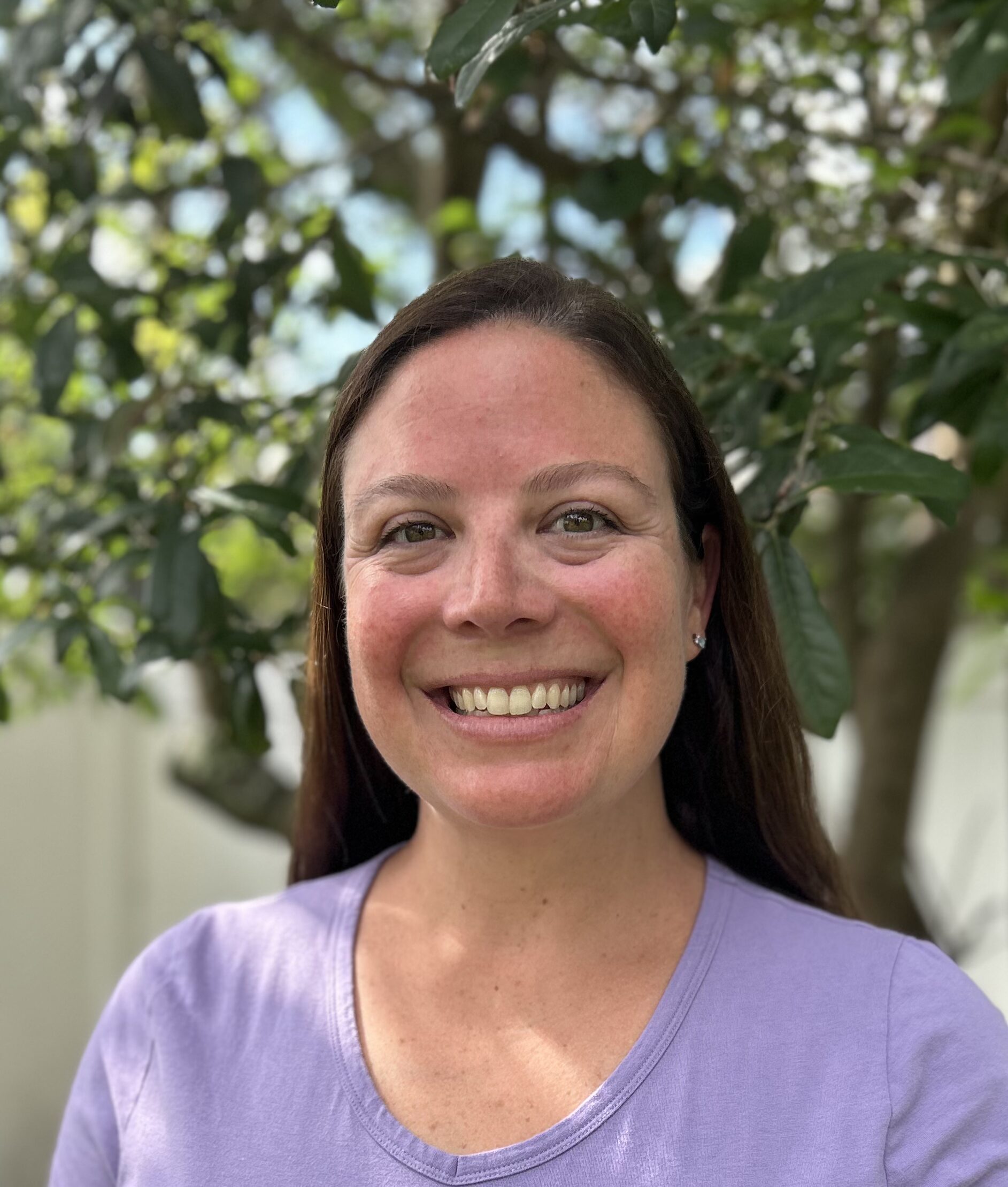 Woman smiling in purple shirt, with brown hair and a tree in the background.
