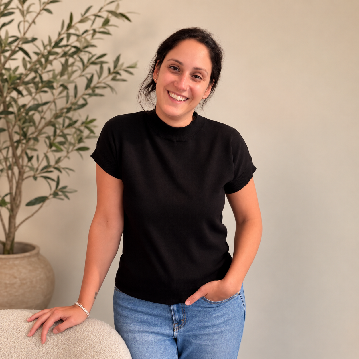 Woman smiling, with hair in a pony tail, one hand in pocket, and one leaning on a table, in a black shirt and jeans, with a potted plant behind her