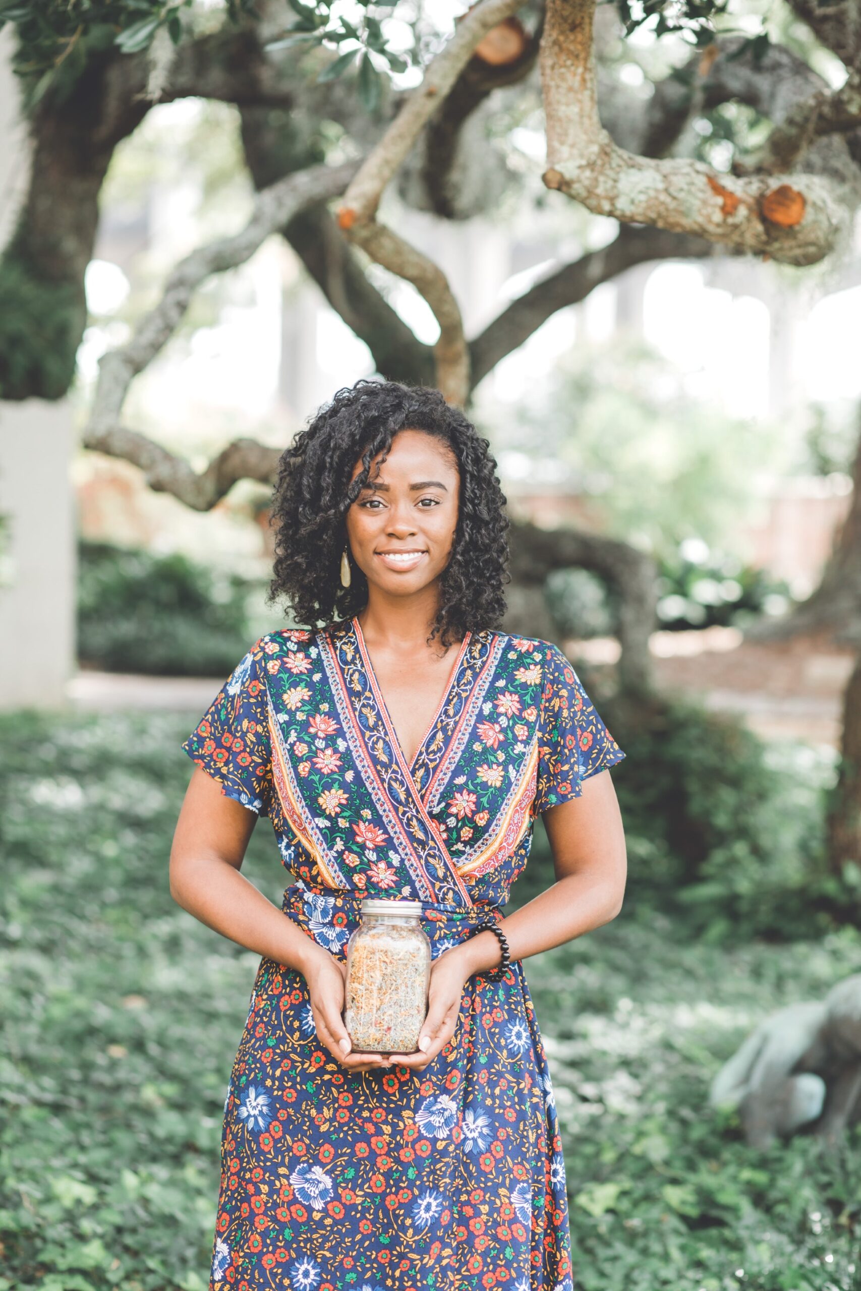 African American Woman with curly black hair smiling, wearing a patterned dress ,holding a glass jar in front of trees.