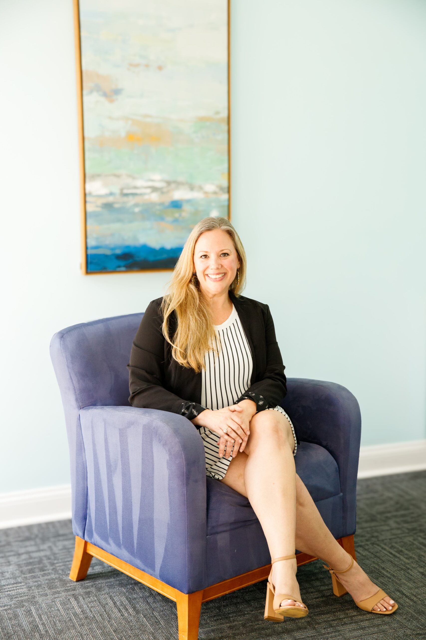 blonde woman sitting in purple chiar smiling in black and white striped dress with paiting behind her